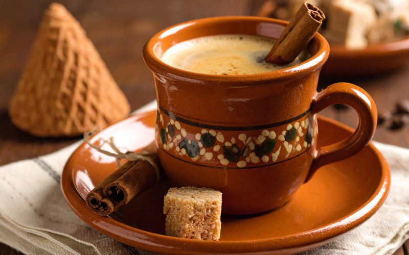 A traditional clay mug filled with steaming Café de Olla, with a cinnamon stick and a cone of piloncillo next to it.